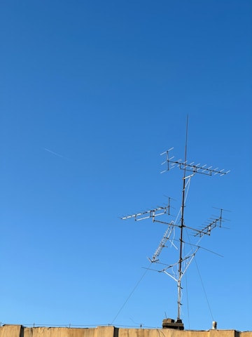 A clear blue sky serves as the backdrop to a rooftop-mounted antenna system, featuring multiple horizontal and vertical elements. The structure is built of metal and stands prominently against the sky. A beige-colored wall is visible at the bottom of the image.