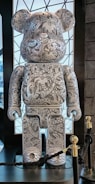 A large, stylized bear figure covered in intricate black-and-white patterns, including abstract shapes, symbols, and doodles. It stands on a platform with a backdrop of a geometric patterned window. The bear features rounded ears and a simplified, blocky form. There are ropes and stanchions around the display.