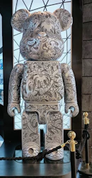 A large, stylized bear figure covered in intricate black-and-white patterns, including abstract shapes, symbols, and doodles. It stands on a platform with a backdrop of a geometric patterned window. The bear features rounded ears and a simplified, blocky form. There are ropes and stanchions around the display.