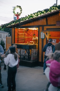 A festive outdoor market stall is adorned with greenery and lights on the roof. Inside, a vendor is preparing food on a grill, with racks behind him containing more food items. People, including children, are gathered around outside, presumably waiting for orders.