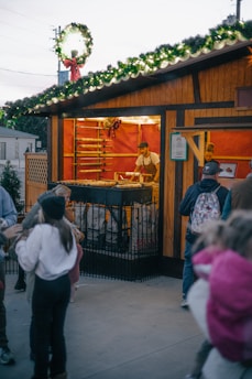A festive outdoor market stall is adorned with greenery and lights on the roof. Inside, a vendor is preparing food on a grill, with racks behind him containing more food items. People, including children, are gathered around outside, presumably waiting for orders.