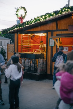 A festive outdoor market stall is adorned with greenery and lights on the roof. Inside, a vendor is preparing food on a grill, with racks behind him containing more food items. People, including children, are gathered around outside, presumably waiting for orders.