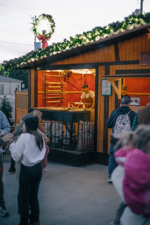 A festive outdoor market stall is adorned with greenery and lights on the roof. Inside, a vendor is preparing food on a grill, with racks behind him containing more food items. People, including children, are gathered around outside, presumably waiting for orders.