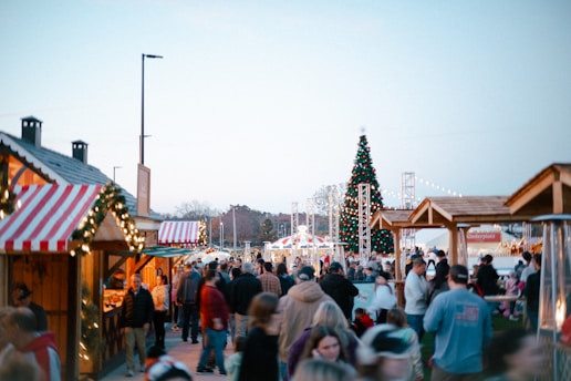 A bustling outdoor market scene with numerous people walking among wooden stalls adorned with holiday decorations. A large Christmas tree, decorated with lights, stands prominently in the background. The atmosphere is lively, with people browsing and socializing.