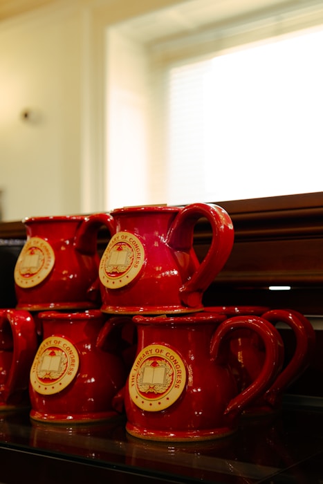 Stacked red coffee mugs with The Library of Congress emblem are displayed on a shelf. The mugs are glossy, with a smooth finish and rounded handles.