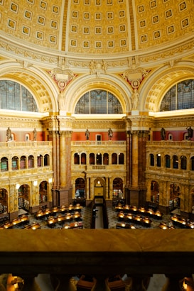 An ornate and grandiose interior of a library with a large, domed ceiling adorned with intricate patterns and golden accents. The architectural design includes large arches, decorative columns, and statues positioned around the upper gallery. The main floor is arranged with multiple reading desks, each illuminated by individual lamps, creating a serene and scholarly atmosphere.
