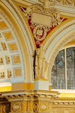 An ornate interior with intricate architectural details, featuring a classical statue in a draped robe. The structure includes decorative moldings and a stained glass window in the background. The color scheme incorporates warm shades of gold and cream, along with a red backdrop adorned with angelic figures and an engraved tablet.