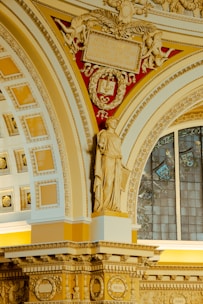 An ornate interior with intricate architectural details, featuring a classical statue in a draped robe. The structure includes decorative moldings and a stained glass window in the background. The color scheme incorporates warm shades of gold and cream, along with a red backdrop adorned with angelic figures and an engraved tablet.