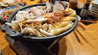 A hot pot filled with various ingredients including prawns, mushrooms, scallops, and other vegetables. The pot is placed on a wooden table with several dishes and bowls in the background, suggesting a dining setting.