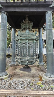 An ornate, ancient-looking lantern is positioned beneath a pavilion with four pillars. The intricate design includes detailed carvings and a decorative top. The base is surrounded by small stones, and the structure is set in a natural, wooded environment.