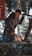 A person in a dimly lit workshop is engaged in blacksmithing. Sparks fly around as they work on a piece of iron on an anvil with a hammer. The person is wearing a dark coat and a beanie, surrounded by various metal tools and equipment, with ornate metalwork in the foreground.