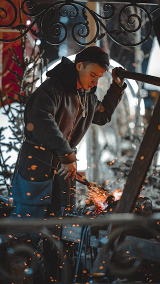 Close-up of a craftsman’s hands shaping a metal piece with a hammer and anvil in a workshop bathed in warm light.