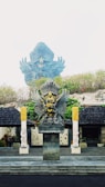 The image features a large, ornate statue of Garuda, a mythical bird-like creature from Hindu and Buddhist mythology, adorned with intricate details and vibrant colors. The Garuda statue is prominently positioned on a stone pedestal, surrounded by greenery and flanked by two smaller statues. The background shows part of a larger Garuda statue, partially obscured by the mist.