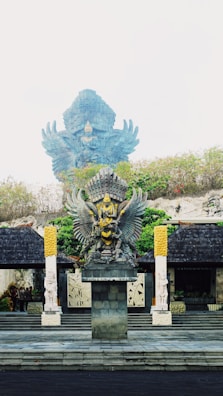 The image features a large, ornate statue of Garuda, a mythical bird-like creature from Hindu and Buddhist mythology, adorned with intricate details and vibrant colors. The Garuda statue is prominently positioned on a stone pedestal, surrounded by greenery and flanked by two smaller statues. The background shows part of a larger Garuda statue, partially obscured by the mist.
