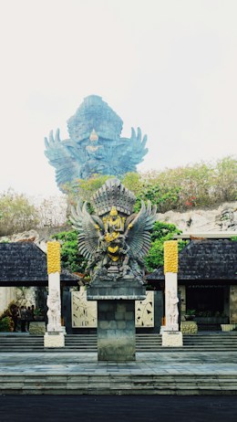 The image features a large, ornate statue of Garuda, a mythical bird-like creature from Hindu and Buddhist mythology, adorned with intricate details and vibrant colors. The Garuda statue is prominently positioned on a stone pedestal, surrounded by greenery and flanked by two smaller statues. The background shows part of a larger Garuda statue, partially obscured by the mist.