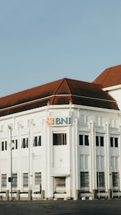 A historic white building with distinct architectural features and a red-tiled roof is prominently displayed. The facade has decorative elements and narrow windows, with a sign featuring the BNI logo. The sky is clear, and the scene is calm.