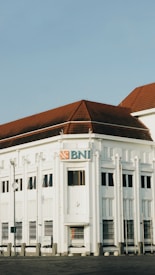 A historic white building with distinct architectural features and a red-tiled roof is prominently displayed. The facade has decorative elements and narrow windows, with a sign featuring the BNI logo. The sky is clear, and the scene is calm.