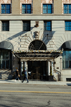 A large, multi-story building with a classic architectural style features multiple windows and symmetrical design elements. The central entrance has a decorative canopy adorned with flowers, and the text 'Hotel Washington' is visible above the door. Two people are walking on the sidewalk in front of the building, one in a black coat and the other in a blue jacket. The weather appears clear with shadows cast on the building façade.