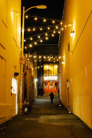 An alleyway illuminated by strings of warm yellow lights hanging overhead, creating a cozy ambiance. The walls on either side are painted in muted colors. At the end of the alley, a lit-up entrance with the word 'Sovereign' is visible, and a person is walking towards it. The scene suggests a quiet urban environment at night.