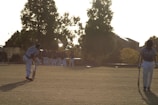 A group of cricket players are visible on a grassy field, surrounded by large trees. Two players in the foreground are walking away from the viewer, holding cricket bats. One player is in a batting pose while the other appears to be walking with a lowered head. The background shows the sun casting long shadows and illuminating the scene.