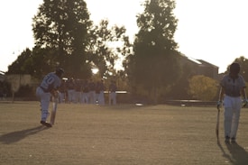 A group of cricket players are visible on a grassy field, surrounded by large trees. Two players in the foreground are walking away from the viewer, holding cricket bats. One player is in a batting pose while the other appears to be walking with a lowered head. The background shows the sun casting long shadows and illuminating the scene.