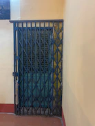 A metal accordion-style security gate is closed in front of a wooden door with a dark, weathered finish. The walls surrounding the door are painted in a pale yellow hue, and a red trim lines the floor. There is a small portion of an artwork visible in the upper left corner.