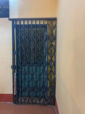 A metal accordion-style security gate is closed in front of a wooden door with a dark, weathered finish. The walls surrounding the door are painted in a pale yellow hue, and a red trim lines the floor. There is a small portion of an artwork visible in the upper left corner.