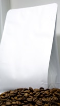 A pile of coffee beans is placed in front of a plain white packaging bag, which appears to be made of paper or plastic. The coffee beans are prominently displayed at the bottom, showing their rich brown color and texture.