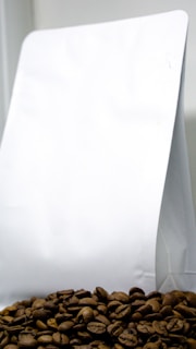 A pile of coffee beans is placed in front of a plain white packaging bag, which appears to be made of paper or plastic. The coffee beans are prominently displayed at the bottom, showing their rich brown color and texture.