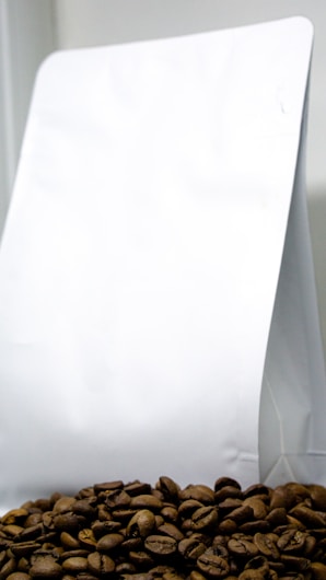 A pile of coffee beans is placed in front of a plain white packaging bag, which appears to be made of paper or plastic. The coffee beans are prominently displayed at the bottom, showing their rich brown color and texture.