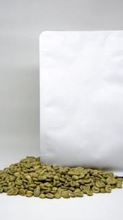 A white coffee bag stands upright with a pile of green coffee beans gathered at its base. The backdrop is a simple and plain white surface, highlighting the natural color of the beans.