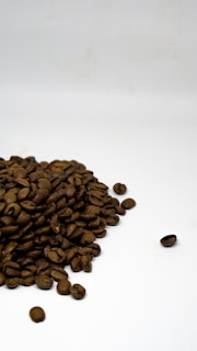 A pile of dark brown coffee beans spread across a plain white surface, with one bean slightly isolated to the right.