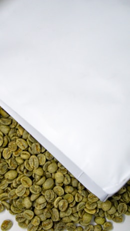A collection of unroasted green coffee beans is partially covered by a white paper or packaging. The beans have a raw, natural appearance and are scattered below the white surface.