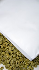 A collection of unroasted green coffee beans is partially covered by a white paper or packaging. The beans have a raw, natural appearance and are scattered below the white surface.