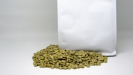 A pile of green coffee beans is placed in front of a large, plain white bag. The beans have an unroasted, raw appearance with a greenish hue.