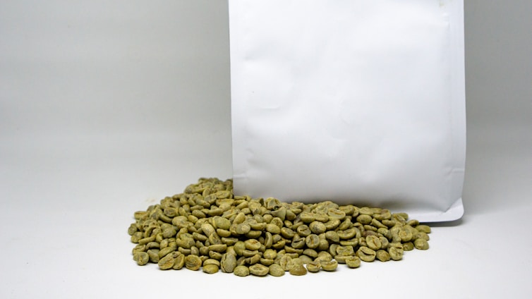 A pile of green coffee beans is placed in front of a large, plain white bag. The beans have an unroasted, raw appearance with a greenish hue.