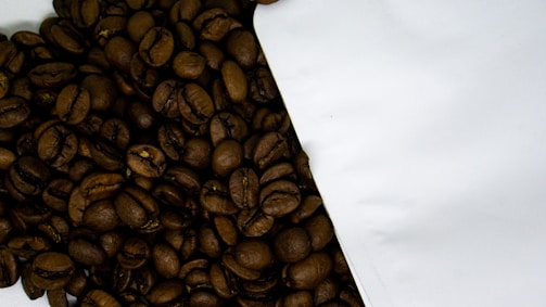 A close-up view of roasted coffee beans spilling out from a white package onto a surface. The coffee beans are dark, shiny, and have a rich texture, indicating freshness.