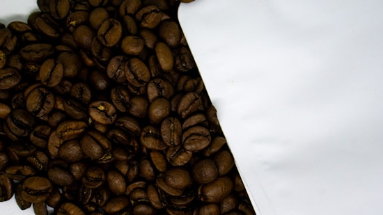 A close-up view of roasted coffee beans spilling out from a white package onto a surface. The coffee beans are dark, shiny, and have a rich texture, indicating freshness.