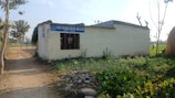 A small, simple concrete building with a corrugated metal roof is situated on a dirt path surrounded by greenery. The building has a sign that reads 'Shree Bhairab Bidhya Niketan' and appears to serve as an educational establishment. There are plants and rocks in the foreground, and leafless trees are scattered around, with open fields extending into the distance.