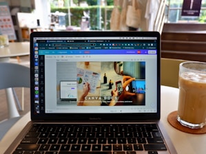 A laptop displaying a Canva webpage open to a design document. The laptop is placed on a table in a cafe setting with a glass of iced coffee on a coaster nearby. The background shows blurred elements of the cafe, including chairs and a window.