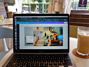 A laptop displaying a Canva webpage open to a design document. The laptop is placed on a table in a cafe setting with a glass of iced coffee on a coaster nearby. The background shows blurred elements of the cafe, including chairs and a window.