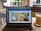 A laptop displaying a Canva webpage open to a design document. The laptop is placed on a table in a cafe setting with a glass of iced coffee on a coaster nearby. The background shows blurred elements of the cafe, including chairs and a window.