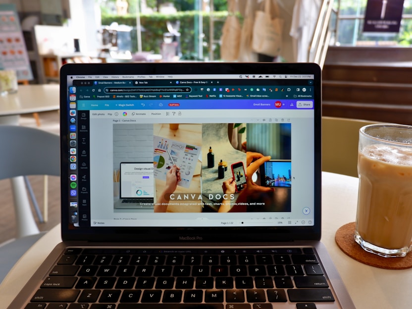 A laptop displaying a Canva webpage open to a design document. The laptop is placed on a table in a cafe setting with a glass of iced coffee on a coaster nearby. The background shows blurred elements of the cafe, including chairs and a window.