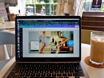A laptop displaying a Canva webpage open to a design document. The laptop is placed on a table in a cafe setting with a glass of iced coffee on a coaster nearby. The background shows blurred elements of the cafe, including chairs and a window.