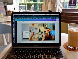A laptop displaying a Canva webpage open to a design document. The laptop is placed on a table in a cafe setting with a glass of iced coffee on a coaster nearby. The background shows blurred elements of the cafe, including chairs and a window.
