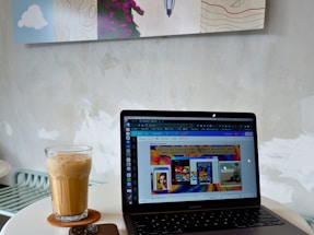 A modern workspace setup featuring a laptop open on a round table. The screen displays a colorful website interface. Beside the laptop is a glass of iced coffee on a coaster and a smartphone. The background reveals a minimalistic wall with abstract art.