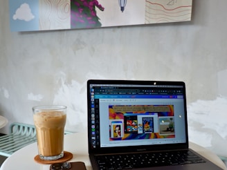 A modern workspace setup featuring a laptop open on a round table. The screen displays a colorful website interface. Beside the laptop is a glass of iced coffee on a coaster and a smartphone. The background reveals a minimalistic wall with abstract art.