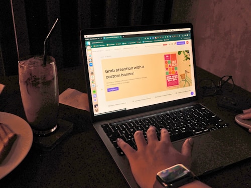 A hand reaches towards a laptop with a website open displaying a banner advertisement. Beside the laptop, a glass with a straw is visible. The scene is dimly lit, possibly in a cafe or home environment. A smartwatch is worn on the wrist of the hand.