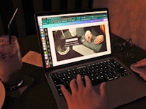 A person is working on a laptop with a design editing software open, displaying a presentation slide featuring two hands typing on a keyboard. The scene is dimly lit, and there is a glass of iced beverage with a straw on the left. A pair of glasses and a mouse are placed beside the laptop.