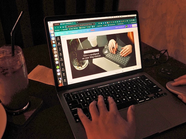 A person is working on a laptop with a design editing software open, displaying a presentation slide featuring two hands typing on a keyboard. The scene is dimly lit, and there is a glass of iced beverage with a straw on the left. A pair of glasses and a mouse are placed beside the laptop.